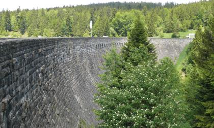 Das Verblendwerk der Schwarzenbachtalsperre aus Raumünzach-Granit Das Foto zeigt eine hohe, im Bogen nach rechts verlaufende steinerne Staumauer aus dunkelgrauem Mauerwerk. Im Vordergrund rechts ein paar Nadelbäume, im Hintergrund bewaldete Höhen.