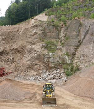 Tiefgründige Vergrusung, Steinbruch Tegernau-Niedertegernau Blick auf eine hohe Steinbruchwand mit bröckeliger Oberfläche links und festen Blöcken rechts im Bild. Im Vordergrund arbeitet ein Radlader.