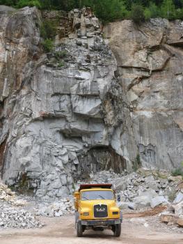 Malsburg-Granit im Steinbruch Malsburg-Marzell Blick auf eine hohe, zerklüftete Steinbruchwand, grau bis hellbraun. Am Fuß der Wand liegen Steinhaufen. Ganz vorne steht ein gelber Lastwagen.