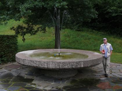„Suppenschüssel“ von Steinklingen aus Heidelberg-Granit Das Bild zeigt einen schüsselförmigen Brunnen aus rosa Gestein. Der Brunnen ist wassergefüllt und in der Mitte befindet sich eine kleine Fontäne. Der Brunnen steht auf einer mit ebenfalls rosa Platten gepflasterten Fläche, im Hintergrund ist Wiese.