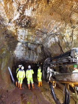 Unter-Tage-Aufnahme aus der Grube Clara mit vererztem Diagonaltrum Blick in das Innere eines Bergwerks. Zu sehen ist ein breiter, gut ausgeleuchteter Stollen mit hoher Decke sowie ein ins Dunkel führender Schacht. Mehrere Personen in gelber Schutzkleidung stehen davor.