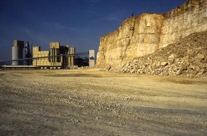 Steinbruch Merdingen mit frisch gesprengtem Haufwerk, im Hintergrund das Kalkwerk Blick auf eine Steinbruchwand aus gelblich braunem Gestein sowie einem Abraumhaufen davor (rechts im Bild). Im Hintergrund links sind Förder- und Verarbeitungsanlagen zu sehen.