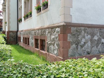 Grundschule in Bötzingen Blick auf die Ecke eines Gebäudes mit heller Fassade und abgesetztem Sockel. Das dunkelgraue Mauerwerk des Sockels ist von rötlichen Steinen eingefasst.