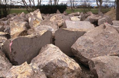 Rohblöcke aus Kaiserstühler Tuffstein im Lager bei Breisach Lager von rötlich grauen, unregelmäßig geformten Gesteinsblöcken. Bei zwei Blöcken im Vordergrund sind glatte, senkrecht verlaufende Schnittkanten sichtbar.
