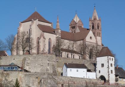 Das Breisacher St. Stephansmünster Blick auf eine Kirche mit hellbraunem Mauerwerk und roten Dächern. Links ist ein breiter, viereckiger Turm erkennbar, rechts zwei unterschiedliche, höhere Türme. Die Kirche thront auf einem Mauerberg. Unten rechts steht ein weißer Torturm.