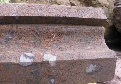 Brauner bis graubrauner Achkarrener Tuffstein in einem Probewerkstück für das Münster in Breisach Das Foto zeigt ein bearbeitetes, längliches Gesteinsstück mit abgerundeten Vertiefungen und geraden Kanten. Die Gesteinsfarbe ist bräunlich grau mit wenigen hellgrauen und zahlreichen dunkelgrauen Flecken.