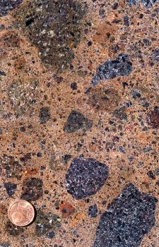 Typischer Kaiserstühler Tuffstein von Achkarren Nahaufnahme einer geschliffenen Gesteinsoberfläche. Grundfarbe rötlich braun, mit größeren Einlässen in dunkelbraun (oben links) und blauschwarz (rechts unten und oben). Eine Münze links unten dient als Größenvergleich.