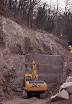 Tuffsteinabbau am Achkarrener Schlossberg Blick auf einen nach rechts abfallenden Waldhang mit offener Seite. Das freiliegende, dunkelgraue Gestein ist in der Bildmitte wie eine Treppe zugeschnitten. Ein gelber Bagger steht davor.