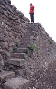 Weinbergsmauer bei Achkarren Das Foto zeigt in Schrägansicht eine hohe Steinmauer, mit Fluchtrichtung rechts. Eine angebaute Steintreppe führt auf die Mauerkrone. Auf der obersten Stufe steht ein Mann mit roter Jacke.