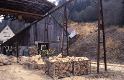 Kalktuffabbau der Fa. Beck bei Bärenthal: Fabrikationshalle Blick auf ein Gesteinslager mit teilweiser Überdachung. Die Gesteinsbrocken liegen in Gitterkörben oder offen herum. Im Hintergrund links ein mit Holz verschaltes Gebäude sowie ein Bagger, rechts ein aufgegrabener Waldhang.