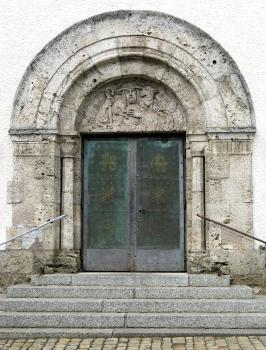 Herz-Jesu-Kirche in Göschweiler: Portal Blick auf einen Kircheneingang mit Steinstufen, Metalltüren und gemauerter, grünlich grauer Umrandung mit aufgesetztem Reliefbogen als Abschluss.
