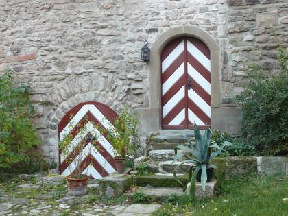 Teilweise aus Kieselsandstein bestehendes Blockmauerwerk im Schloss Honhardt bei Crailsheim Blick auf graues Mauerwerk und zwei Eingänge eines burgähnlichen Gebäudes. Die Eingangstüren sind mit rot-weißem Pfeilmuster bemalt; die linke liegt tiefer, zur rechten Tür führt eine kleine Steintreppe hinauf.