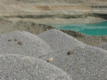 Kieshalden Im Vordergrund sind mehrere graue Kieshalden zu sehen. Dahinter erblickt man einen See sowie die hellbraunen Abbauwände einer Kiesgrube.