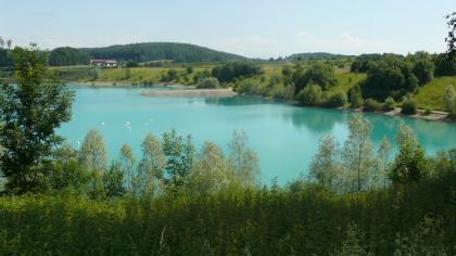 Baggersee in einer Kiesgrube bei Pfullendorf Blick über Baumwipfel auf einen hellblauen See und die angrenzende, von Bäumen bestandene Uferböschung. Im Hintergrund ragt ein aufgeschütteter Dammweg in den See. Auf dem See schwimmen Schwäne.
