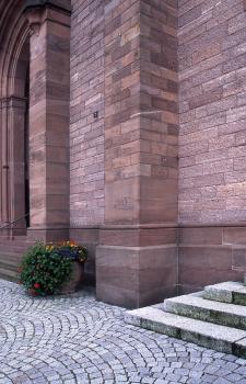 Die Basilika St. Bonifazius, u. a. aus Leisberg-Porphyr errichtet Teilansicht einer Kirchenwand aus rötlich grauem Mauerwerk mit Stützpfeilern und Eingangsbereich links hinten. Der Vorplatz ist gepflastert.