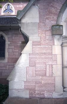 Die St. Johanniskirche, u. a. aus Leisberg-Porphyr errichtet Teilansicht einer Kirchenfassade aus rötlich grauem Mauerwerk. Zu sehen ist rechts ein Vorbau mit Pfeiler und Türbogen; an den Rändern mit helleren Steinen abgesetzt. Links oben ist ein Ornament-Fenster erkennbar.