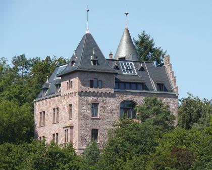 Villa Stroh auf dem Cäcilienberg Blick auf eine von Bäumen umstandene Villa aus rötlich grauem Mauerwerk. Das Gebäude hat zwei Türme, einen Treppengiebel, blaue und silbergraue Dächer, mehrere Dachgauben und Dachfenster.