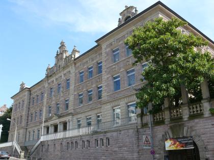 Leisberg-Porphyr an der Vincenti-Grundschule in Baden-Baden Blick auf ein mehrstöckiges, langgezogenes Schulgebäude aus rötlich grauem Mauerwerk. Zu sehen sind Eckteile und Mittelteil. Im Mittelteil befindet sich der Eingang, mit Säulen flankiert. Davor verläuft ein V-förmiger Treppenaufgang.