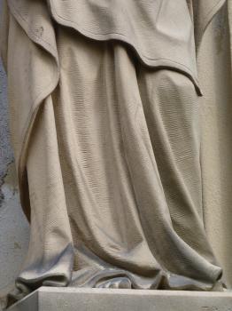 Ausschnitt aus einer Grabsteinplastik auf dem Alten Friedhof in Freiburg-Herdern, Lettenkeuper-Sandstein Teilansicht einer aus bräunlich grauem Stein gehauenen Figur mit faltenreichem, bis auf die Füße reichendem Gewand.