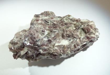 Lithium-reicher Pegmatit Nahaufnahme eines Handstückes von Mineralgestein vor hellem Hintergrund. Der Gesteinsbrocken zeigt verschiedene Farbnuancen, von grau über gelbgrün bis rosaviolett. Auch weißliche Flecken sind erkennbar.