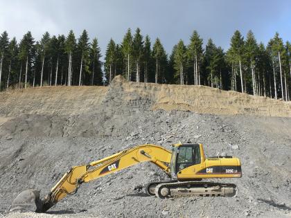 Gewinnung von Opalinuston aus dem Mittleren Jura in der Tongrube Schömberg / Withau Blick auf die leicht gerundete, oben mit Nadelbäumen besetzte Wand eines Steinbruches. Das Gestein ist oben bräunlich, nach unten hin grau. Im Vordergrund ist ein abgestellter gelber Bagger zu sehen.