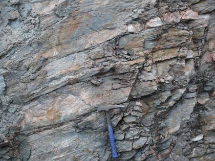 Biotitreicher Paragneis aus dem Steinbruch Freiamt-Keppenbach Nahaufnahme von feinscherbigem, rechts blättrigem Gestein, das in dünnen Lagen wie Buchseiten aufeinanderliegt. Links sind zudem die Farben heller, rechts dunkler. Ein Hammer unten am Bildrand zeigt die Größenverhältnisse an.