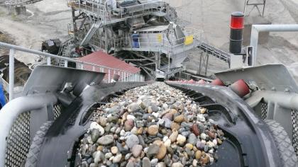 Kiese und Sande im Nassabbau Blick von oben auf ein Förderband und eine Fördermaschine. Auf dem Förderband liegen zahlreiche kleine und größere Steine.