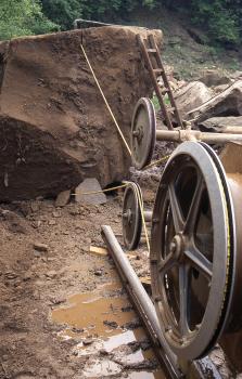 Gewinnung von Niederhofener Sandstein, Zerteilung großer Blöcke durch die Seilsäge Das Foto zeigt die Zerteilung eines großen braunen Steinblockes mit Hilfe einer Seilsäge. Rechts vorne ein großes Antriebsrad der Säge, im Hintergrund links der Steinblock. Dazwischen die Seile der Säge sowie zwei kleine Laufrollen.