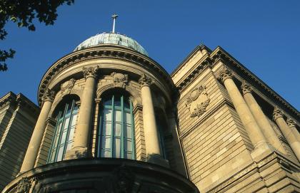 „Königliche Centralstelle für Gewerbe und Handel“ in Stuttgart aus Heilbronner Sandstein, heutiges Haus der Wirtschaft Teilansicht vom obersten Stockwerk eines Gebäudes mit gelblich braunem Mauerwerk. Im Bild ist ein runder, turmartiger Erker sowie rechts ein eckiger Anbau mit Säulen um die Fenster.
