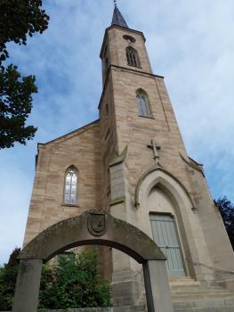 Ev. Kirche in Waldangelloch aus Weiler Sandstein Aufwärts fotografierter Kirchturm aus hellem Mauerwerk mit Eingangsportal rechts und seitlichem Anbau links. Im Vordergrund ein steinerner Wappenbogen.