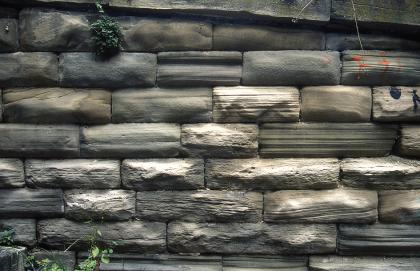 Treppenmauer an der Karlshöhe aus Stuttgarter Schilfsandstein Teilansicht einer Steinmauer mit dunkelgrauen bis graubraunen Mauersteinen, zum Teil mit weißlichen Verfärbungen.