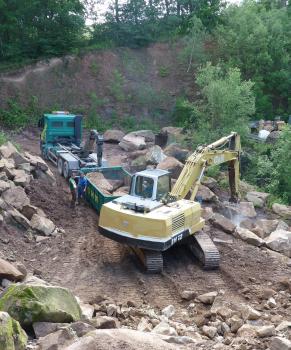 Gewinnung von Niederhofener Sandstein, Sortierung der Blöcke nach Größe Blick von oben in einen Steinbruch. Ein gelber Bagger sortiert und verlädt größere Steinblöcke in einen bereitstehenden Container. Im Hintergrund erhebt sich ein brauner, von Bäumen und Sträuchern bewachsener Hügel.