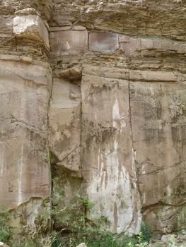 Blick auf eine hohe Steinbruchwand. Im oberen Teil befinden sich dünnplattige Abraumschichten, getrennt durch größere Blöcke. Darunter steht eine von senkrechten Rissen durchzogene Kluftwand an.