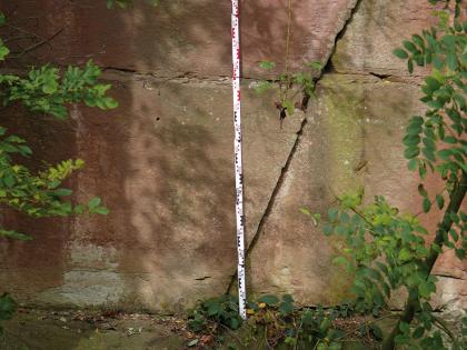 Roter Schilfsandstein im auflässigen Steinbruch bei Winnenden-Hertmannsweiler Nahaufnahme von rötlich braunem Gestein, oben und unten von feinen Rissen durchzogen. Rechts der Bildmitte verläuft ein stärkerer, schräg-senkrechter Riss. In der Bildmitte ist eine Meßlatte an den Stein gelehnt.