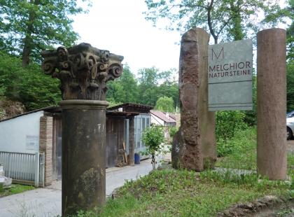 Zufahrt zum Werk der Fa. Melchior im alten Freudentaler Steinbruch Das Foto zeigt den Eingangsbereich eines Werkes für Natursteine. Rechts säumen drei unterschiedliche gestaltete, rötlich bis schwärzlich graue Steinsäulen die Zufahrt.