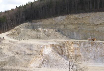 Hochreine Kalksteine aus dem Steinbruch Blaubeuren-Altental Blick auf eine abgestufte, nach links abfallende hellgraue bis bräunliche Steinbruchwand mit Abraumhaufen und einem Fahrweg. Die gerundete Kuppe ist mit Nadelbäumen bewachsen.