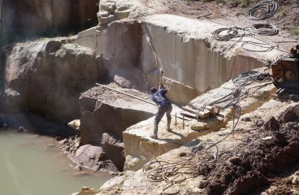 Abbau im Stubensandsteinbruch Pliezhausen-Rübgarten Blick von oben in einen Steinbruch. Ein auf einem Steinblock stehender Arbeiter versucht, mit einem Hammer Keile in das Gestein zu schlagen. Im Hintergrund Steinbruchwände. Links unten steht Wasser.