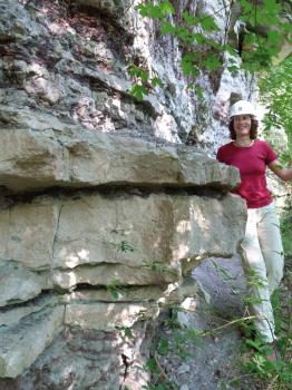 Stubensandstein in einem alten Steinbruch bei Herrenberg-Kayh Seitlicher Blick auf eine von Bäumen beschattete Gesteinswand. Zwei Drittel des Gesteins ist großbankig und bräunlich, darüber zeigen sich rötlich graue, streifige und knollige Stellen. Rechts steht eine Frau mit Schutzhelm.