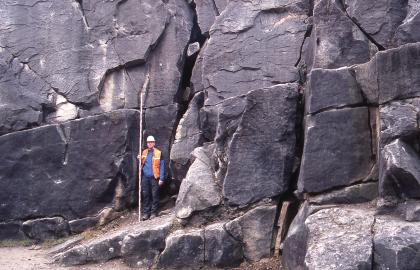 Auflässiger Steinbruch am Sandacker südlich von Stetten Blick auf eine alte Steinbruchwand mit violettgrauen, massigen Blöcken. Das Gestein ist von zahlreichen Rissen und senkrechten Klüften durchzogen. Links der Bildmitte hält ein Mann in Schutzkleidung eine Messlatte an das Gestein.