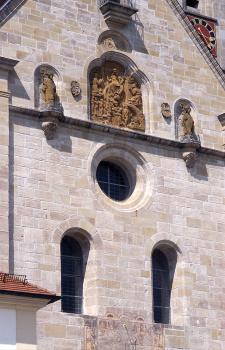Teil der Südfassade der Basilika St. Vitus in Ellwangen Teilansicht einer Kirchenfassade aus grauem bis hellbraunem Mauerwerk. Zu sehen sind Fenster mit runden Bögen, ein Rundfenster, Figuren und ein bronzefarbenes Relief oberhalb der Bildmitte.