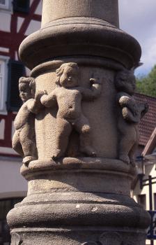 Brunnensäule des Marktbrunnens in Oberndorf am Neckar aus Stubensandstein Teilansicht einer aus rötlich grauem Stein gefertigten Säule. In der Bildmitte reihen sich mehrere kleine Steinfiguren um die Säule.