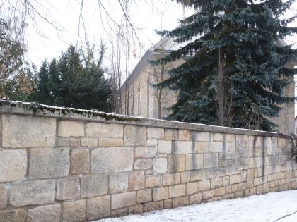 Friedhofsmauer und ev. Kirche Dettenhausen Blick auf eine hellbraune bis hellgraue Steinmauer. Im Hintergrund, hinter der Mauer und einem Nadelbaum, steht ein Kirchengebäude aus ähnlich hellem Stein.