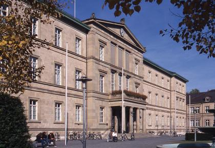 Neue Aula der Universität Tübingen aus Stubensandstein und Schilfsandstein Seitlicher Blick auf ein großes Gebäude aus rötlich grauem Mauerwerk. Der etwas vorstehende Mittelteil des Gebäudes hat einen von Säulen flankierten, überdachten Eingang.