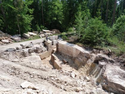 Probeabbau 2012 im Waldgebiet Oberer Neubronn Blick von oben in ein flaches Steinbruchgelände, umgeben von Wald. Erkennbar sind Fahrwege, eine niedrige, über Eck gehende und abgestufte Gesteinswand an der Waldgrenze (rechts im Bild) sowie lose Steinblöcke (links).