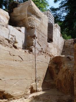 4 m tiefer Probeabbau im Waldgebiet Oberer Neubronn Blick von unten auf eine hochragende, hellbraune Steinbruchwand. Links oben hat die Wand ausgebrochene Stellen. Rechts schließen sich stufige, im Schatten liegende Gesteinswände an.