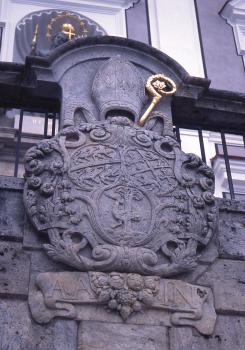 Bischöfliches Wappen am Konventgebäude der Abtei Neresheim, Oberjura-Dolomit Nahaufnahme eines aus violettgrauem Stein gearbeiteten Wappens. Im oberen Teil ist eine Bischofsmütze sowie ein vergoldeter Stab erkennbar. Das Wappen ist Teil einer Gebäudefassade oder einer davor angebrachten Mauer.