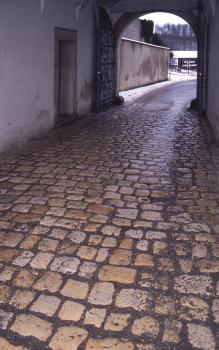 Pflaster aus Oberjura-Kalkstein, Abtei Neresheim Blick auf bräunliches bis graues Kopfsteinpflaster, das zwischen Gebäudewänden auf einen bogenförmigen Durchgang rechts oben zuführt.