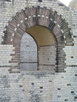 Stützmauer eines römischen Amphitheaters bei Basel aus Oberjura-Kalkstein Teilansicht einer älteren, hellgrauen Steinmauer. In der Mitte der Mauer ist ein bogenförmiger, offener Durchlass, was die große Dicke der Mauer erkennbar macht. Der Bogen des Durchlasses wird durch dunklere Steine betont.