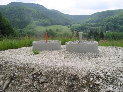 Inklinometer- und Grundwassermessstelle in der Rutschmasse Das Bild zeigt zwei Betonröhren, die in einen flachen Erd- und Kieshügel eingelassen sind. Aus den Betonröhren ragen dünne Messröhren heraus. Im Hintergrund bewaldete Hügel und Berge.