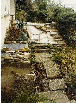 Zerstörte Gartenterrasse Mittig befinden sich einige Bodenplatten, die chaotisch zueinander verschoben sind. Außenrum Büsche und links eine Hauswand.